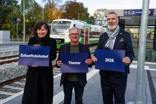 Drei Personen stehen am Bahnhof Themar und "2026" in die Kamerahalten Schilder mit der Aufschrift "Zukunftsbahnhof", "Themar", 